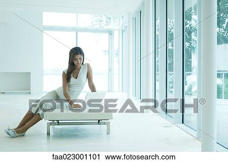Young woman sitting on settee, using laptop computer, looking down, full length View Large Photo Image Stock Image - Young woman sitting on settee, using laptop computer, looking down, full length. Fotosearch
