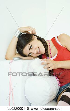 Stock Image - Young woman sitting on sofa, leaning head on arm, smiling at camera. Fotosearch