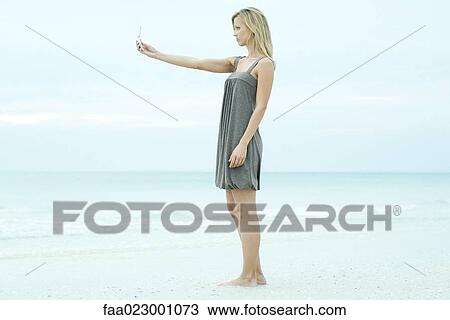 Young woman standing at the beach, photographing self with cell phone, side view View Large Photo Image Stock Image - Young woman standing at the beach, photographing self with cell phone, side view. Fotosearch