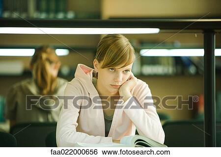 Stock Photograph - Young woman studying in university library, holding head, looking away. Fotosearch