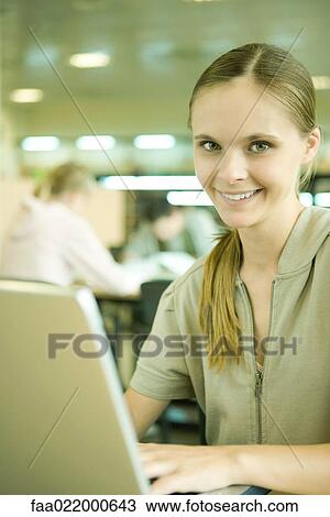Young woman using laptop in library, smiling at camera View Large Photo Image Stock Image - Young woman using laptop in library, smiling at camera. Fotosearch
