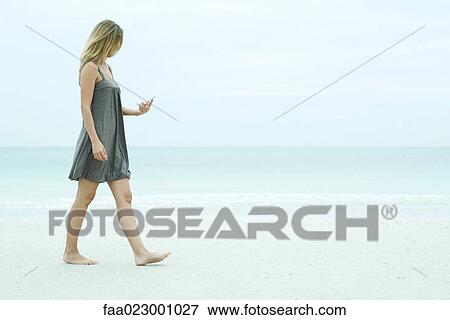 Stock Photo - Young woman walking on the beach, looking at cell phone. Fotosearch