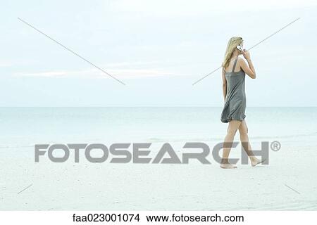 Young woman walking on the beach, using cell phone, full length View Large Photo Image Picture - Young woman walking on the beach, using cell phone, full length. Fotosearch