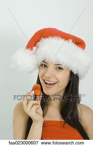 Young woman wearing Santa hat, holding up strawberry, smiling at camera, portrait View Large Photo Image Stock Photo - Young woman wearing Santa hat, holding up strawberry, smiling at camera, portrait. Fotosearch