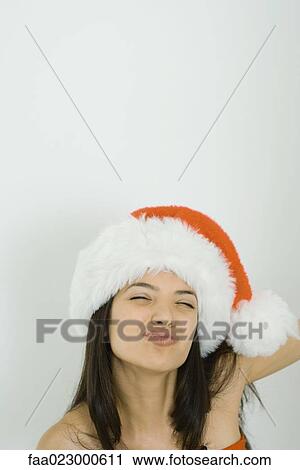 Stock Image - Young woman wearing Santa hat, puckering lips, squinting, portrait. Fotosearch