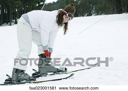 Stock Image - Young woman wearing skis, bending over to adjust boots. Fotosearch