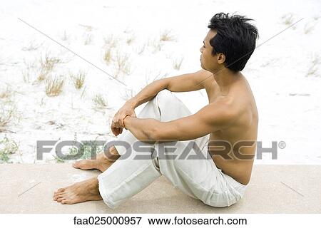 Stock Photo - Barechested man sitting near dune, full length. Fotosearch