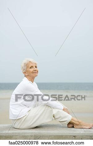 Barefoot senior woman sitting on low wall, smiling, beach in background, full length View Large Photo Image Stock Photo - Barefoot senior woman sitting on low wall, smiling, beach in background, full length. Fotosearch