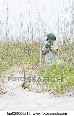 Stock Photograph - Boy crouching in tall grass, holding toy gun, looking at camera. Fotosearch
