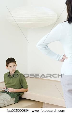 Boy holding video game glaring at mother who watches him with hands on hips View Large Photo Image Picture - Boy holding video game glaring at mother who watches him with hands on hips. Fotosearch
