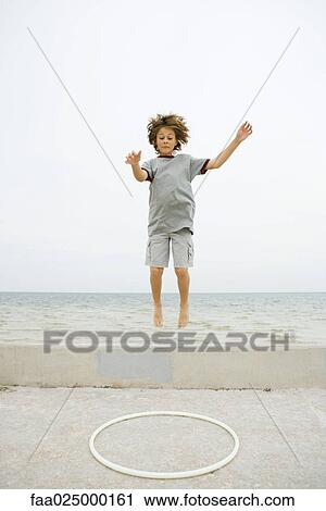 Stock Image - Boy jumping off low wall toward plastic hoop, arms raised. Fotosearch