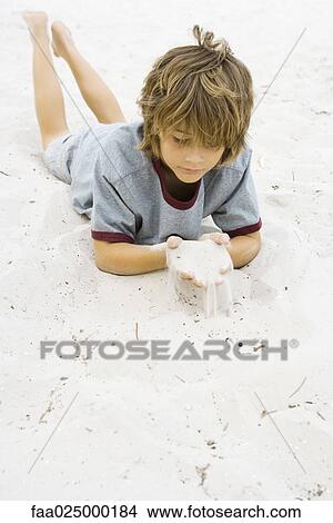 Boy lying on the ground, looking down at handful of sand View Large Photo Image Picture - Boy lying on the ground, looking down at handful of sand. Fotosearch