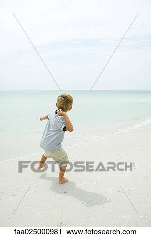 Stock Image - Boy on beach throwing unseen object toward ocean, rear view, full length. Fotosearch