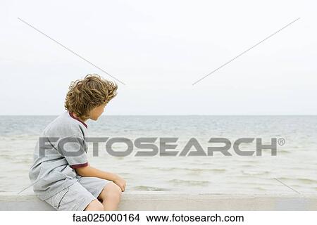 Boy sitting on low wall at the beach, looking at view View Large Photo Image Picture - Boy sitting on low wall at the beach, looking at view. Fotosearch