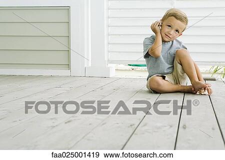 Stock Photo - Boy sitting on the ground listening to seashell, smiling. Fotosearch