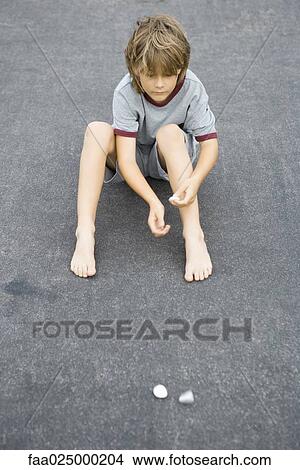 Boy sitting on the ground throwing pebbles, high angle view View Large Photo Image Picture - Boy sitting on the ground throwing pebbles, high angle view. Fotosearch