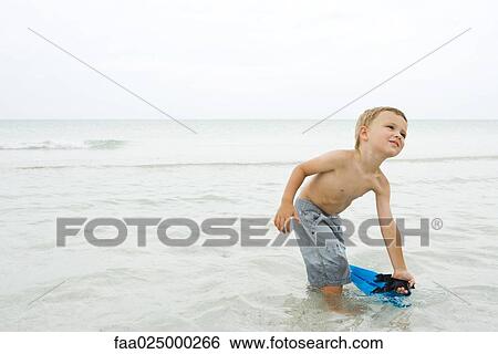 Stock Photograph - Boy standing knee deep in water, picking up flippers, looking up. Fotosearch