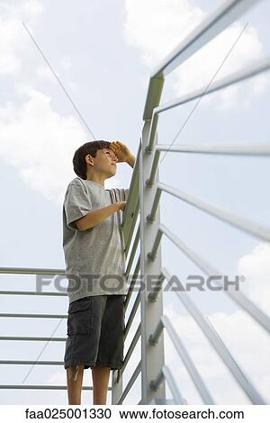 Stock Image - Boy standing on balcony, looking at view, low angle view. Fotosearch