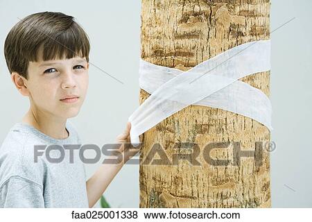 Stock Photo - Boy wrapping bandage around tree trunk, looking at camera. Fotosearch