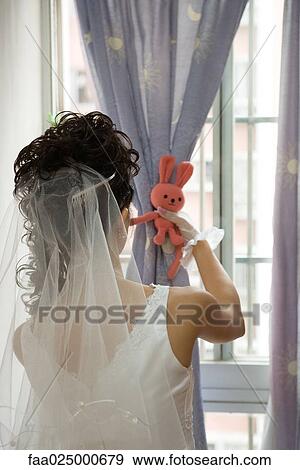 Stock Photo - Bride facing window, tying curtain with stuffed toy, rear view. Fotosearch