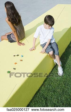 Stock Photo - Brother and sister sitting back to back on low wall next to toys, high angle view. Fotosearch