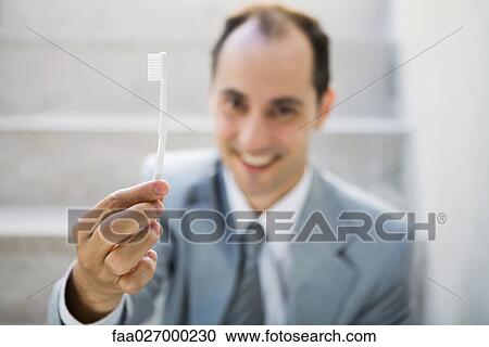 Stock Image - Businessman holding up toothbrush, smiling at camera, focus on foreground. Fotosearch