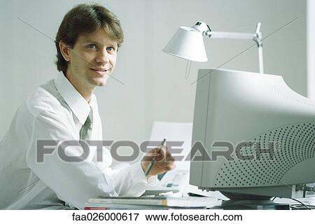 Businessman sitting at desk in front of computer, smiling, looking up View Large Photo Image Stock Photo - Businessman sitting at desk in front of computer, smiling, looking up. Fotosearch