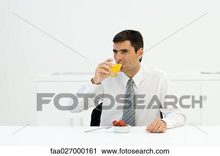 Stock Image - Businessman sitting at table, drinking orange juice, looking away. Fotosearch