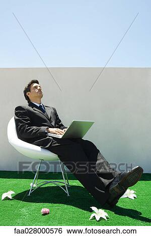Businessman sitting in chair on artificial turf, using computer, full length View Large Photo Image Stock Photograph - Businessman sitting in chair on artificial turf, using computer, full length. Fotosearch