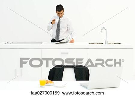 Businessman standing in kitchen, reading newspaper and drinking coffee, breakfast on table in foreground View Large Photo Image Stock Photo - Businessman standing in kitchen, reading newspaper and drinking coffee, breakfast on table in foreground. Fotosearch