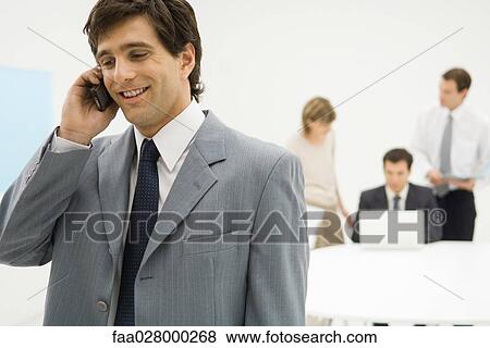 Businessman using cell phone, smiling, colleagues in background View Large Photo Image Stock Photo - Businessman using cell phone, smiling, colleagues in background. Fotosearch