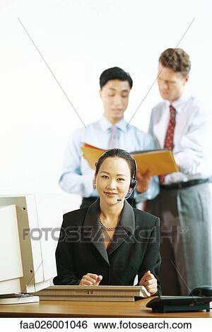 Stock Photograph - Businesswoman sitting at desk, wearing headset, smiling at camera, male colleagues standing in background. Fotosearch