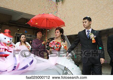 Stock Image - Chinese wedding, bride and groom leaving under confetti, bride covered by red parasol, decorated car in foreground. Fotosearch