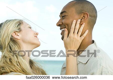 Stock Photograph - Couple smiling at each other, woman's hand on man's cheek, side view. Fotosearch