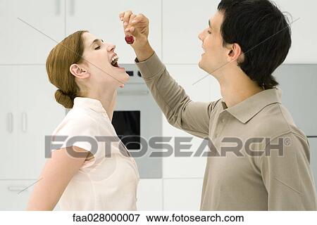 Couple standing together in kitchen, man feeding woman cherries, side view View Large Photo Image Stock Photo - Couple standing together in kitchen, man feeding woman cherries, side view. Fotosearch