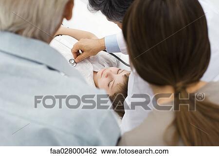 Stock Image - Doctor listening to boy's heart with stethoscope while family members watch, over the shoulder view. Fotosearch