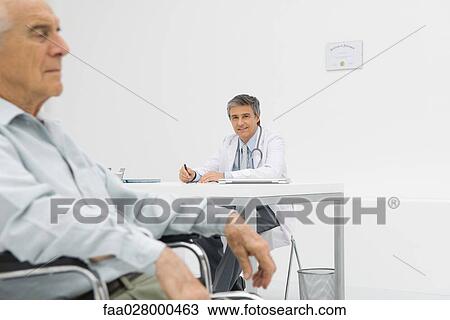 Doctor writing at desk in office, smiling at camera, patient sitting in wheelchair in foreground View Large Photo Image Stock Image - Doctor writing at desk in office, smiling at camera, patient sitting in wheelchair in foreground. Fotosearch