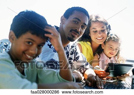 Family sitting together at table outdoors, father and son smiling at camera, mother and daughter looking at menu View Large Photo Image Stock Photograph - Family sitting together at table outdoors, father and son smiling at camera, mother and daughter looking at menu. Fotosearch