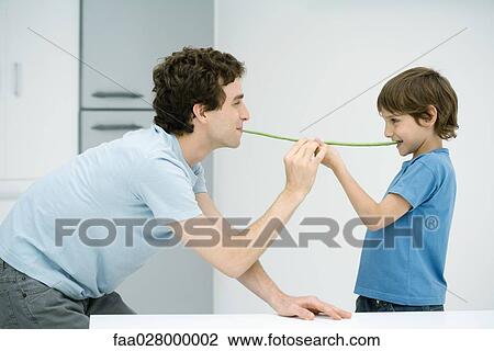Stock Image - Father and son standing face to face, holding up green beans between their mouths, side view. Fotosearch
