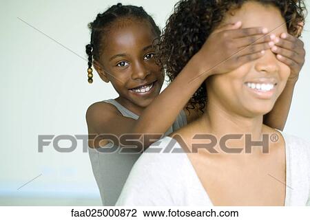 Stock Image - Girl placing hands over her mother's eyes from behind, smiling at camera. Fotosearch