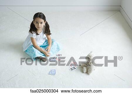 Girl sitting on the ground playing card game with teddy bear, looking up at camera View Large Photo Image Picture - Girl sitting on the ground playing card game with teddy bear, looking up at camera. Fotosearch