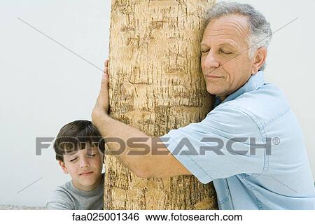 Stock Photograph - Grandfather and grandson hugging tree trunk, eyes closed. Fotosearch