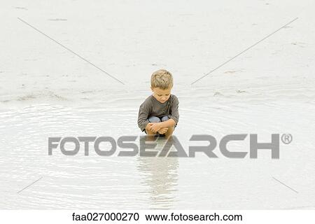 Little boy crouching in water at the beach, looking down View Large Photo Image Stock Image - Little boy crouching in water at the beach, looking down. Fotosearch