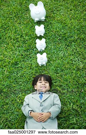 Little boy in full suit lying on grass, line of piggy banks above head View Large Photo Image Stock Photo - Little boy in full suit lying on grass, line of piggy banks above head. Fotosearch