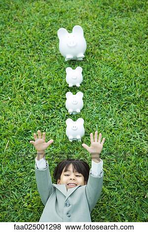 Stock Photo - Little boy in full suit lying on grass, line of piggy banks above head. Fotosearch