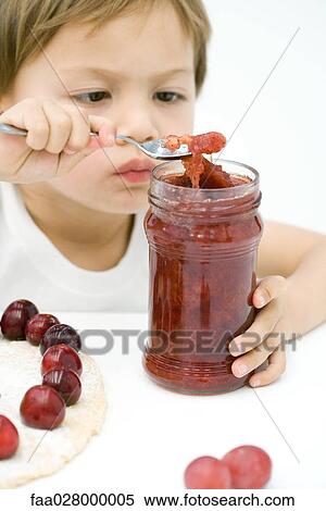 Stock Photography - Little boy scooping jam out of jar with spoon, tart and cherries on table in front of him. Fotosearch