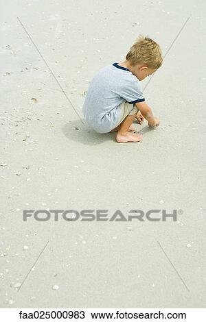 Stock Image - Little boy squatting on beach picking up seashells, full length. Fotosearch