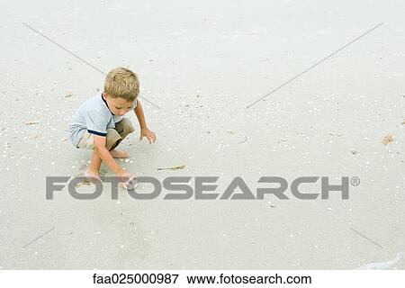 Stock Photo - Little boy squatting, picking up ball on beach, full length. Fotosearch