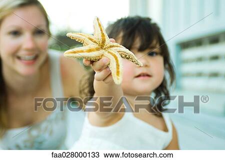 Little girl holding up starfish, looking at camera, mother smiling in background View Large Photo Image Stock Image - Little girl holding up starfish, looking at camera, mother smiling in background. Fotosearch