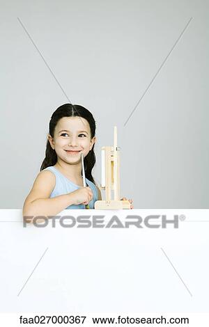 Stock Photo - Little girl in front of easel, holding paintbrush, looking away, smiling. Fotosearch